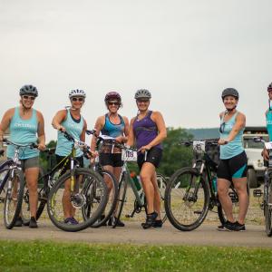 Group of cyclists, 5 women and 1 man, smile for the camera while standing alongside their bikes